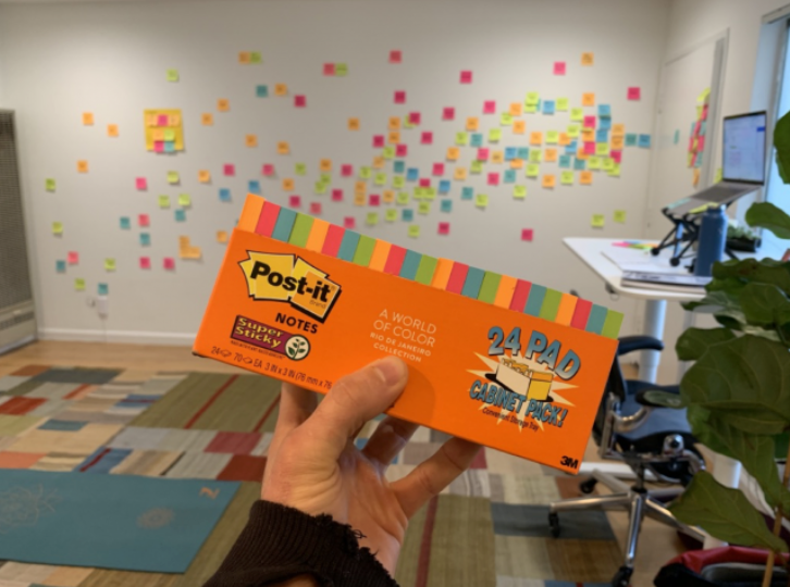 Hands holding a refill pack of Post-it notes in front of a wall covered in colorful stickies forming a storyboard.