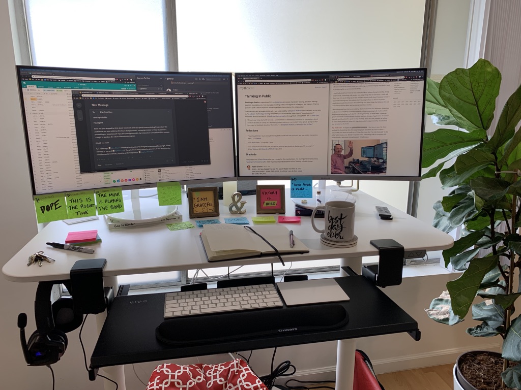 Brian's standing desk with a curved dual-monitor setup: Slack and notifications on the left, the Thinking In Public memo on mythos.one on the right, with handwritten stickies framing the workspace.