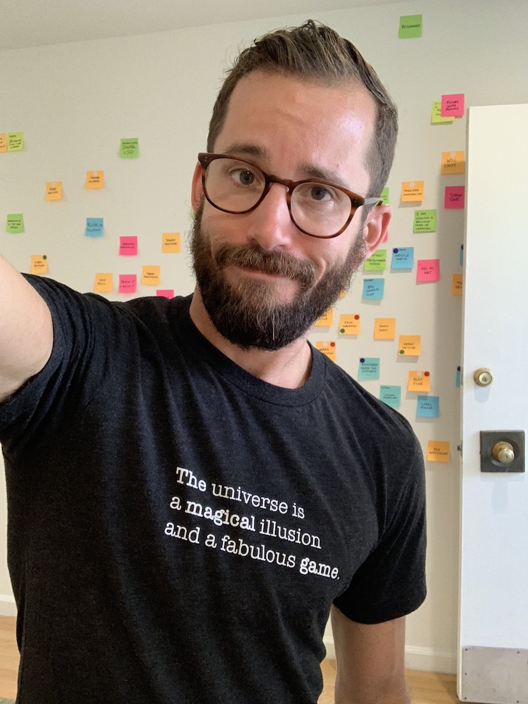 Brian wearing a black t-shirt that reads 'The universe is a magical illusion and a fabulous game,' a selfie taken in front of his colorful sticky-note wall.