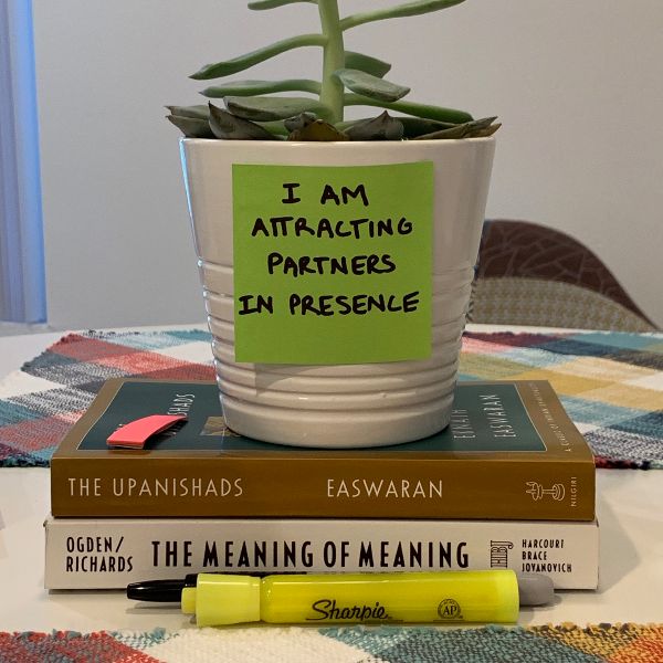 A green sticky note reading 'I AM ATTRACTING PARTNERS IN PRESENCE' stuck to a small succulent pot, atop The Upanishads and The Meaning of Meaning, with a yellow Sharpie in the foreground.